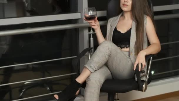 Vacker flicka med kaukasiska utseende med långt hår sitter vid panoramafönstret läser en bok som håller ett glas rött vin och njuter av natten staden — Stockvideo