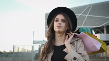 Gözlüklü, yağmurluklu ve şapkalı güzel bir kadın otoparkta kameraya poz veren samimi bir gülümseme ile modern bir binanın arka planında alışveriş yaparken hediye çantaları tutuyor.