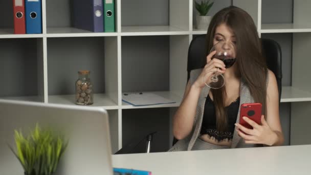 Joven director guapo de la compañía sentado en la oficina sosteniendo un teléfono en sus manos y bebiendo una copa de vino tinto — Vídeo de stock