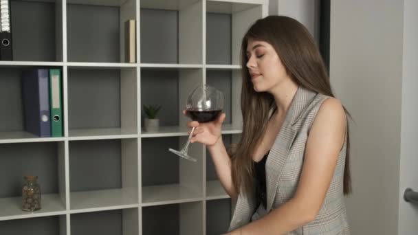 Ung vacker företagsledare sitter på kontoret på kanten av bordet hålla en telefon i händerna och dricka ett glas rött vin efter en hårt arbetande produktiv dag — Stockvideo