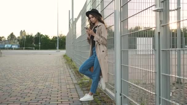 Bella ragazza in piedi per strada con un telefono in mano. Bruna con i capelli lunghi in un cappello nero e mantello marrone — Video Stock