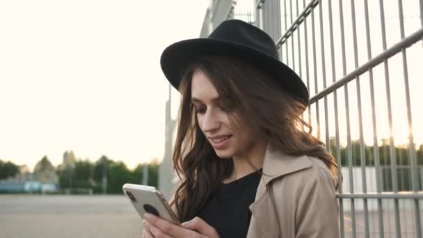 Bella ragazza in piedi per strada con un telefono in mano. Bruna con i capelli lunghi in un cappello nero e mantello marrone — Video Stock