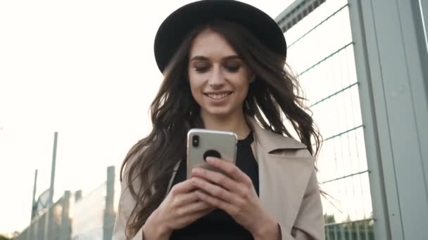 Красивая девушка ходит по улице с телефоном в руках и переписывается в социальных сетях. Брюнетка с длинными волосами в черной шляпе и коричневом плаще — стоковое видео