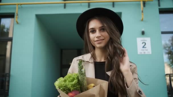 Hermosa mujer con estilo en una capa marrón y sombrero negro camina por la calle cerca de edificios modernos, sosteniendo un paquete de verduras y frutas y un teléfono en sus manos. Entrega de alimentos — Vídeo de stock