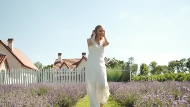 Καυκάσια 25χρονη όμορφη ξανθιά με μακρύ λευκό φόρεμα περπατάει σε ένα χωράφι με λεβάντα σε ένα ηλιόλουστο καλοκαιρινό βράδυ. Πολλά μοβ λουλούδια. — Αρχείο Βίντεο