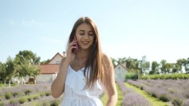 Beyaz elbiseli, uzun saçlı ve samimi bir gülümsemesi olan güzel esmer kız lavanta tarlasında yürüyor ve telefonda konuşuyor. Mor çiçekler. Doğa