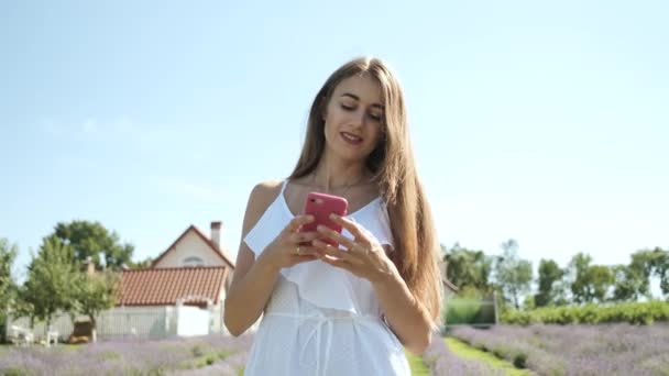 Vacker flicka med kaukasiskt utseende i en vit klänning med långt hår och ett uppriktigt leende går på ett lavendelfält och tittar på telefonen. Lila blommor. Natur — Stockvideo