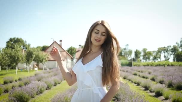 Bella ragazza bruna in un abito bianco con i capelli lunghi e un sorriso sincero cammina su un campo di lavanda. Fiori viola. Natura — Video Stock