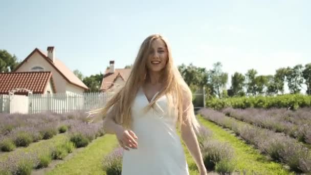 Blanke 25-jarige mooie blonde in een lange witte jurk loopt op een lavendelveld in een zomerse zonnige avond. Veel paarse bloemen — Stockvideo