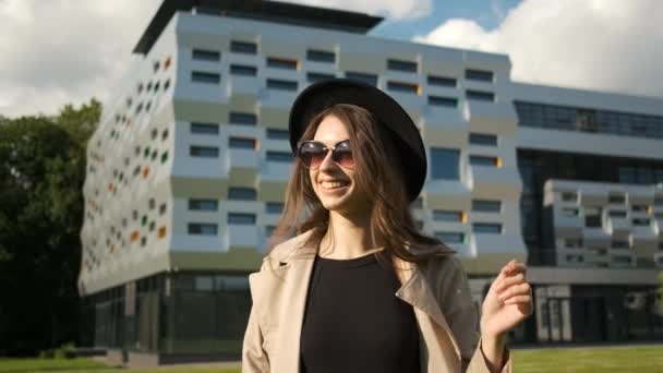 Mooi meisje loopt door de straat in de buurt van een mooi business center. Brunette met lang haar in een zwarte hoed en bruine mantel — Stockvideo