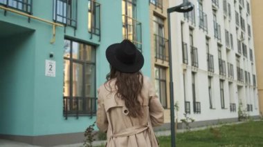 Kahverengi pelerinli, siyah şapkalı güzel bir kadın elinde meyve ve sebze paketiyle modern binaların yakınındaki caddede yürüyor.