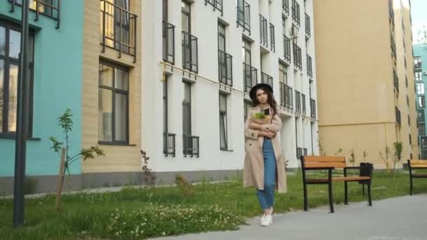 Hermosa mujer con estilo en una capa marrón y sombrero negro camina por la calle cerca de edificios modernos, sosteniendo un paquete de frutas y verduras Entrega de alimentos — Vídeos de Stock