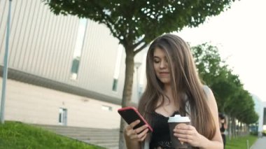 Kafkas görünümlü şık gri takım elbiseli güzel bir iş kadını öğle yemeği molasında ofisin yanında yürüyor kahve ya da çay içiyor ve telefona bakıyor.