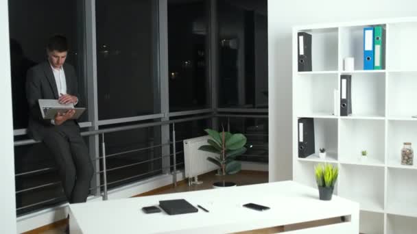 Director of the company in a suit stands in his office near the panoramic window overlooking the night city and solves work issues working on a laptop — Stock Video