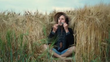 Sakallı ve hasır şapkalı vahşi bir adam buğdayın üzerinde oturmuş kadim bir enstrüman çalıyor. Müzisyen