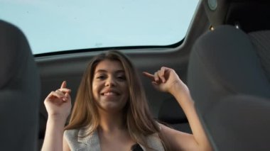 Gri takım elbiseli güzel bir iş kadını panoramik çatılı bir arabanın arka koltuğunda oturur başarılı bir projeden sonra müzik dinler ve dans eder.