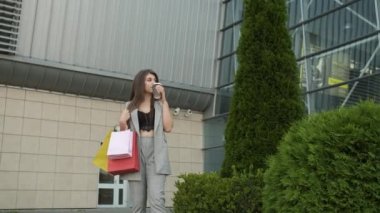 Tarz sahibi gri takım elbiseli ve yakası açık bir kadın alışverişten dönüyor ve kahve ve çay içiyor. Hediye paketleri. Modern binalar