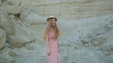 Güzel bir model kum ocağının yanında yürüyor ve kameraya poz veriyor. Kanyon. Sarı şapkalı, pembe elbiseli sarışın kız.