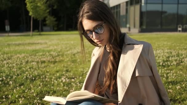 Стильна дівчина в окулярах і коричневий плащ сидить в парку на траві і читає книгу і посміхається в камеру. Подарункові пакети. Сонячний літній вечір — стокове відео