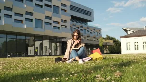 Gözlüklü ve kahverengi pelerinli şık bir kız parkta çimlerin üzerinde oturur ve bir kitap okur ve kameraya gülümser. Hediye paketleri. Güneşli yaz akşamı — Stok video