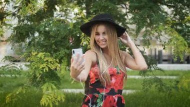 25 yaşında güzel bir sarışın, beyaz tenli, samimi bir gülüşü, uzun bir elbisesi ve siyah bir şapkası var ve bir bankta oturuyor. Neşeli bir kadın müzik dinler ve telefonda fotoğraf çeker.