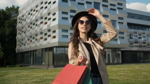 Fille dans un imperméable brun et porte un chapeau noir tout en marchant dans le parc. Retourne à la maison avec des paquets cadeaux shopping — Video