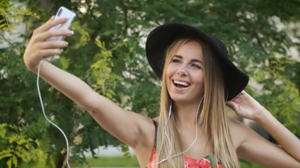 Красивая 25-летняя блондинка кавказской внешности с искренней улыбкой в длинном платье и черной шляпе, сидящая на скамейке в парке. Женщина весело слушает музыку и фотографирует по телефону. — стоковое видео
