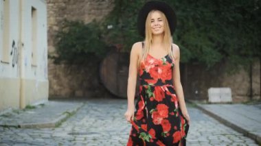 Beyaz görünümlü, samimi bir gülüşü, güzel uzun bir elbisesi ve siyah şapkası olan güzel bir sarışın sabah şehrinde dolaşıyor. Kameranın önünde poz veren bir turist. Model