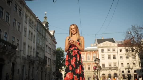 Blond caucasian woman walks walks around the morning city in a long dress and holds a phone in her hand — Stock Video