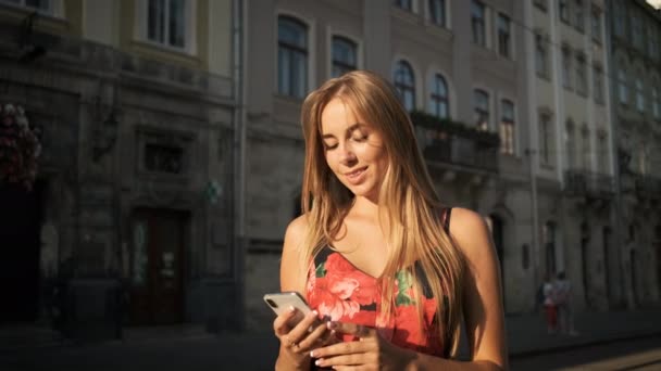 Blond caucasian woman walks walks around the morning city in a long dress and holds a phone in her hand — Stock Video