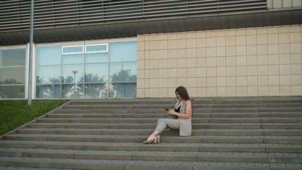 Vacker affärskvinna i en snygg grå kostym av kaukasiskt utseende sitter på gatan på trappan nära kontoret under en lunchrast, njuter av en kopp kaffe eller te och tittar på telefonen — Stockvideo
