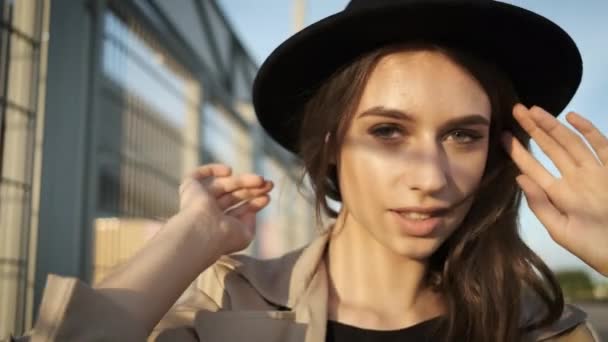 Όμορφο κορίτσι μοντέλο στέκεται και βάζει σε ένα καπέλο στο δρόμο. Μελαχρινή με μακριά μαλλιά σε μαύρο καπέλο και καφέ μανδύα. Κορνίζα από πίσω — Αρχείο Βίντεο
