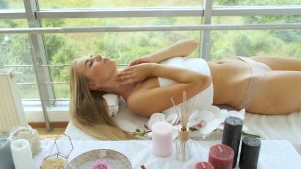 Una mujer rubia de aspecto caucásico se encuentra en un spa con ventanas panorámicas y velas y flores sobre la mesa. Relajación y salud — Vídeo de stock