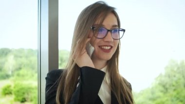 Gözlüklü ve şık takım elbiseli bir kadın, şirket yöneticisi panoramik pencerenin yanındaki ofiste duruyor ve müşterilerle telefonda konuşuyor. İnsanlarla iletişim