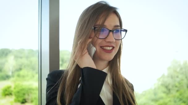 Egy szemüveges nő és egy elegáns öltöny, a cégvezető áll az irodában, közel a panorámaablakhoz, és telefonon beszél az ügyfelekkel. Kommunikáció emberekkel — Stock videók
