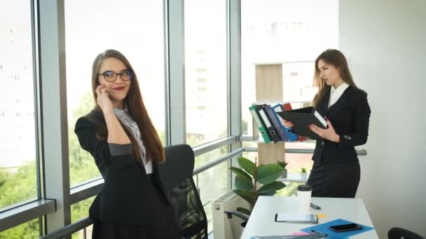 Een vrouw met een bril en een stijlvol pak, de bedrijfsleider staat in het kantoor in de buurt van het panoramische raam en praat aan de telefoon met klanten. Communicatie met mensen — Stockvideo
