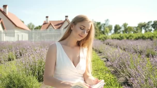 25-летняя блондинка сидит на лавандовом поле, читает книгу и расслабляется — стоковое видео