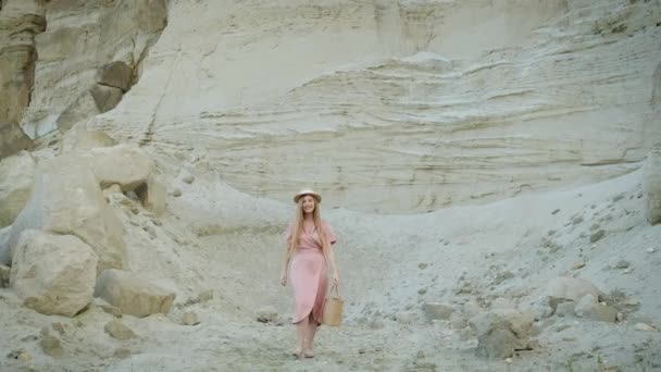 美しいですモデルブロンドでピンクのドレスとわら帽子歩みでザ砂の峡谷でハンドバッグで彼女の手とポーズのためにカメラスローアップ彼女の帽子 — ストック動画