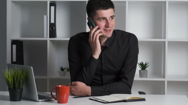 Een lachende bedrijfsadviseur in een zwart shirt zit aan een bureau in het kantoor en praat met klanten — Stockvideo