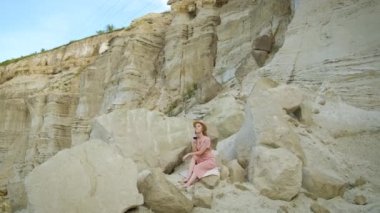 Kafkas görünümlü sarı şapkalı bir kız bir taş ocağında bir taşın üzerinde oturup bir bardak kırmızı şarap içiyor. Rahatla.