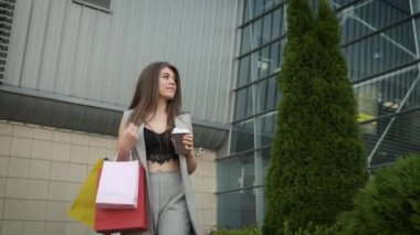 Tarz sahibi gri takım elbiseli ve yakası açık bir kadın alışverişten dönüyor ve kahve ve çay içiyor. Hediye paketleri. Modern binalar