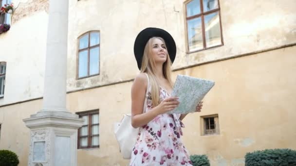 Woman tourist blonde caucasian appearance walks around the morning city in a long dress and hat holds a map and explores the city — Stock Video