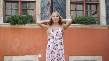 Elbise giymiş sarışın bir manken bir Avrupa şehrinde dolaşıyor ve kameraya poz veriyor. Güzel gülümseme