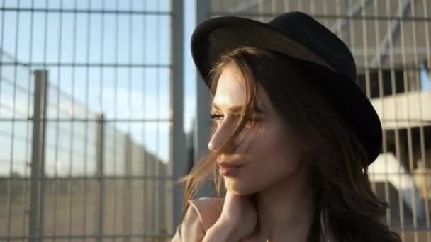 Modelo menina bonita fica e coloca um chapéu na rua. Morena com cabelos longos em um chapéu preto e capa marrom. Moldura de retrato da parte de trás — Vídeo de Stock