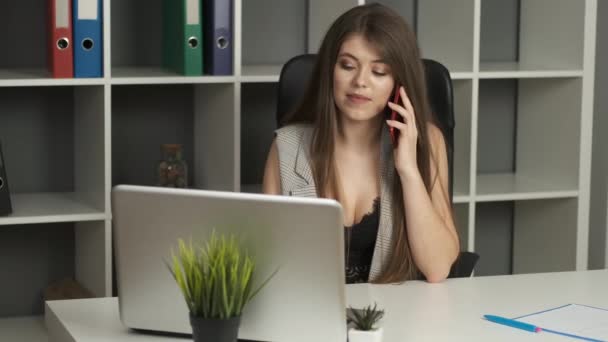 Joven mujer de negocios hermosa de apariencia caucásica con ojos marrones trabaja en su escritorio, sosteniendo un teléfono en sus manos hablando con los clientes — Vídeo de stock