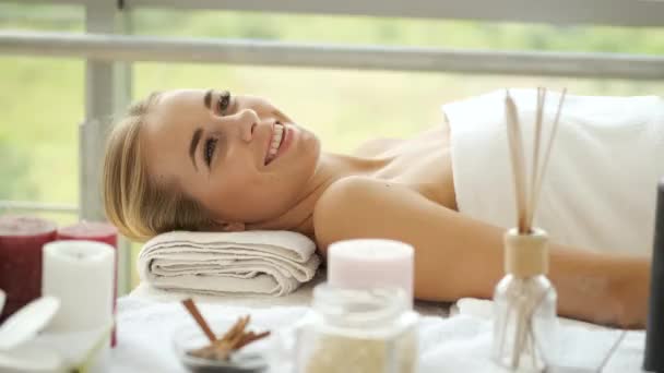 A blonde Caucasian-looking woman lies in a spa with panoramic windows and candles and flowers on the table. Relaxation and health — Stock Video