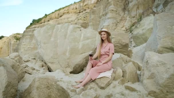A girl in a hat of Caucasian appearance blond hat sits on a stone in a sand quarry and drinks a glass of red wine. Relax — Stock Video
