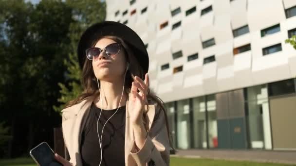 Mooie stijlvolle vrouw in bril, regenjas en hoed luistert actief naar muziek op hoofdtelefoon en spinnen. Wandelingen in het park op de achtergrond van moderne gebouwen — Stockvideo