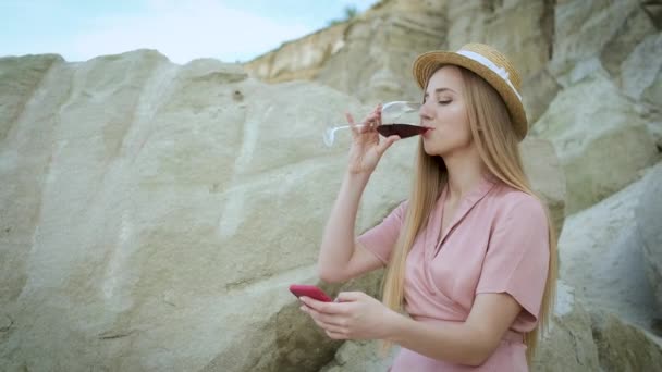 Ragazza turistica di 25 anni con un cappello biondo sguardo caucasico seduta su una roccia in una cava di sabbia e bere un bicchiere di vino rosso e tenere in mano un telefono — Video Stock