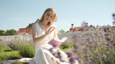 25 yaşında sarışın bir kadın lavanta tarlasında oturmuş kitap okuyor.